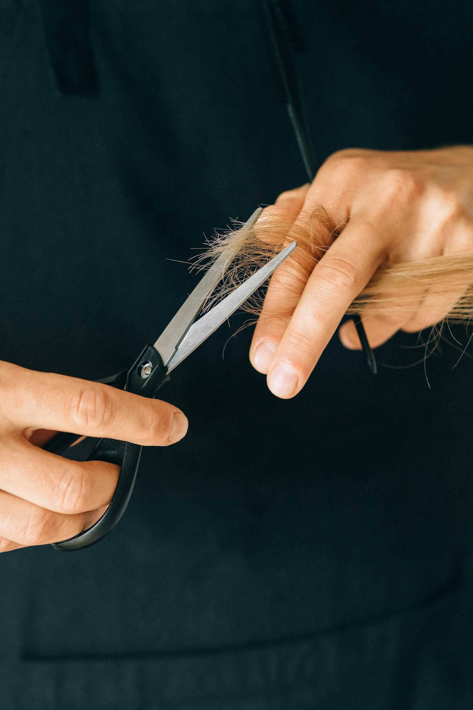 Hands cutting hair with scissors against a dark background.
